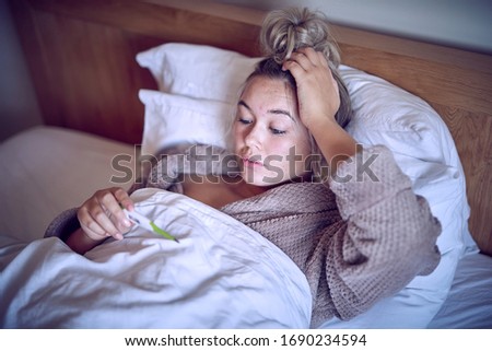 Woman with flu in bed as she blows her nose and looks at a thermometer to check her temperature 