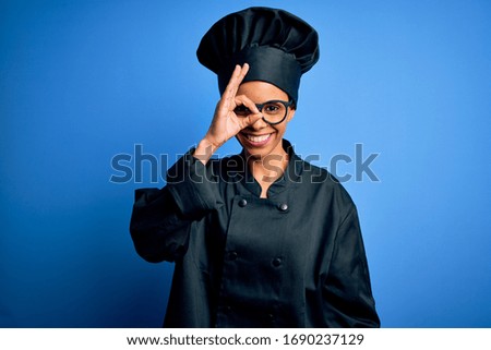Young african american chef woman wearing cooker uniform and hat over blue background doing ok gesture with hand smiling, eye looking through fingers with happy face.