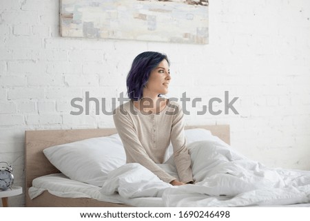 Attractive woman with colorful hair looking away and smiling on bed in bedroom