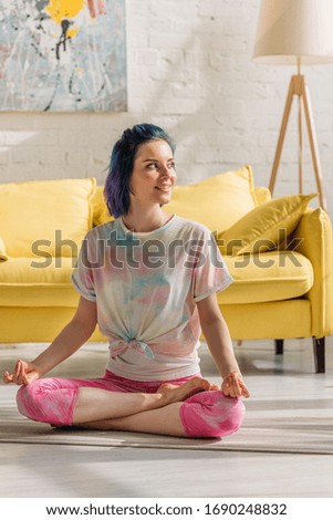 Girl with colorful hair smiling, looking away and meditating on yoga mat in living room