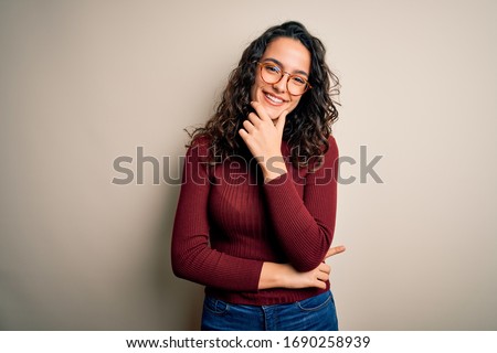 Beautiful woman with curly hair wearing casual sweater and glasses over white background looking confident at the camera smiling with crossed arms and hand raised on chin. Thinking positive.