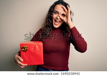 Young beautiful woman with curly hair holding valentine gift over isolated white background with happy face smiling doing ok sign with hand on eye looking through fingers