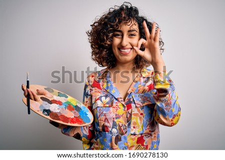 Young beautiful curly arab artist woman painting using brush and palette with colors doing ok sign with fingers, excellent symbol