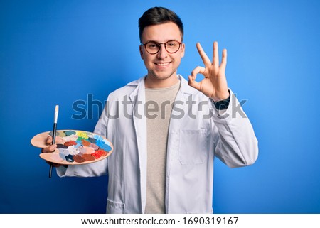 Young handsome caucasian artist man holding paintbrush and painter palette doing ok sign with fingers, excellent symbol
