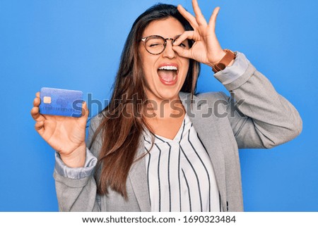 Young hispanic business woman holding credit card over blue isolated background with happy face smiling doing ok sign with hand on eye looking through fingers