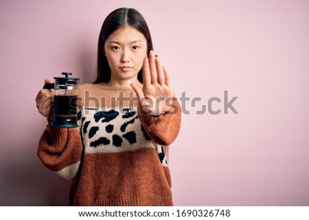 Young asian woman making a glass of coffe using french press coffee maker over pink background with open hand doing stop sign with serious and confident expression, defense gesture