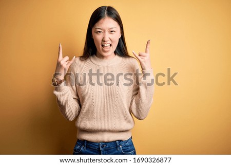 Young beautiful asian woman wearing casual sweater over yellow isolated background shouting with crazy expression doing rock symbol with hands up. Music star. Heavy music concept.