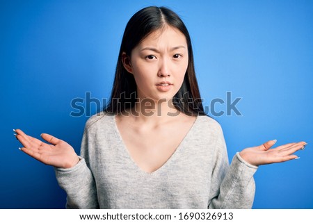 Young beautiful asian woman wearing casual sweater standing over blue isolated background clueless and confused with open arms, no idea concept.