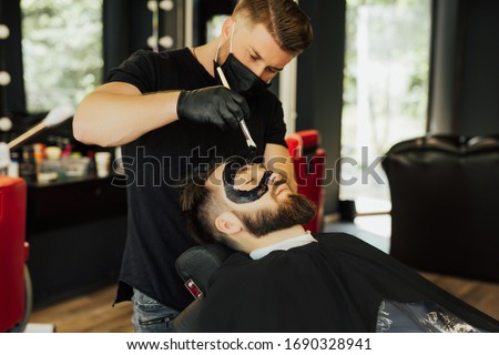 Barber applies black charcoal mask to man face to clean pore skin and remove acne from nose in barber shop. Spa and skin care for men. Young male lying on chair in barber shop. Royalty-Free Stock Photo #1690328941