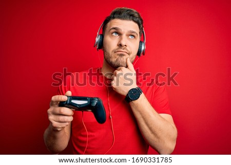 Young gamer man with blue eyes playing video games using gamepad joystick serious face thinking about question, very confused idea