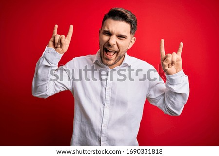 Young business man with blue eyes wearing elegant shirt standing over red isolated background shouting with crazy expression doing rock symbol with hands up. Music star. Heavy music concept.