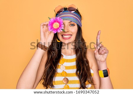 Young beautiful hippie woman with blue eyes wearing sunnglasses holding pink flower over eye surprised with an idea or question pointing finger with happy face, number one