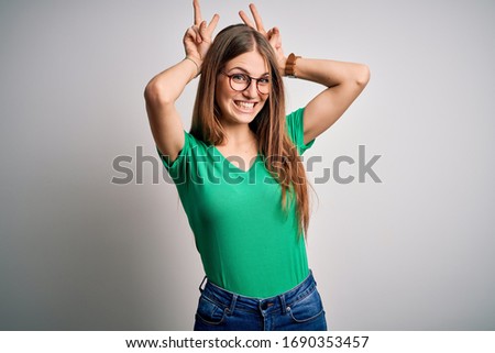 Young beautiful redhead woman wearing casual green t-shirt and glasses over white background Posing funny and crazy with fingers on head as bunny ears, smiling cheerful