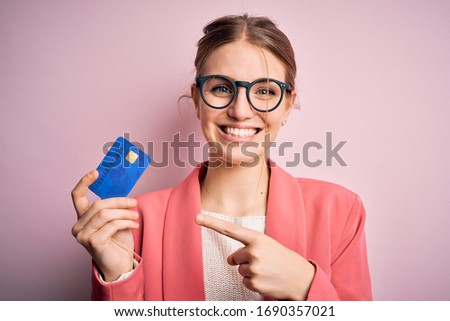 Young beautiful redhead woman holding credit card over isolated pink background very happy pointing with hand and finger