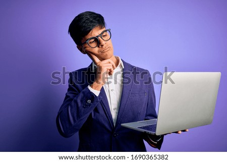 Young handsome business man wearing glasses working using laptop over purple background serious face thinking about question, very confused idea