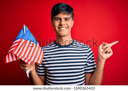 Young handsome patriotic man holding united states flags celebrating independence day very happy pointing with hand and finger to the side
