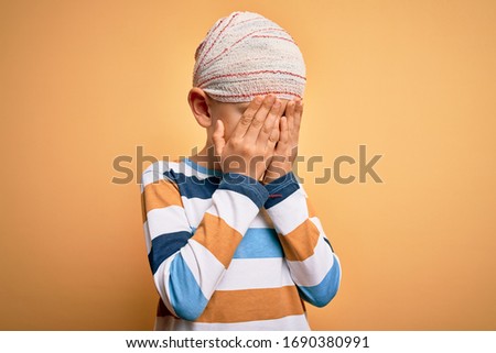 Young little caucasian kid injured wearing medical bandage on head over yellow background with sad expression covering face with hands while crying. Depression concept.
