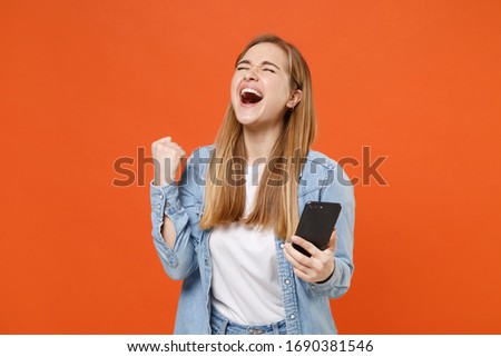 Overjoyed young woman girl in casual denim clothes posing isolated on orange wall background studio portrait. People lifestyle concept. Mock up copy space. Using mobile phone, doing winner gesture