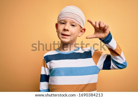 Young little caucasian kid injured wearing medical bandage on head over yellow background smiling and confident gesturing with hand doing small size sign with fingers looking and the camera. Measure