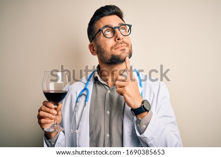 Young doctor man wearing stethoscope drinking a glass of fresh wine over isolated background serious face thinking about question, very confused idea