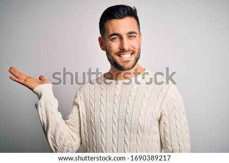 Young handsome man wearing casual sweater standing over isolated white background smiling cheerful presenting and pointing with palm of hand looking at the camera.