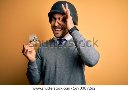 Handsome businessman wearing military helmet holding reminder paper with war message with happy face smiling doing ok sign with hand on eye looking through fingers