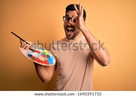 Young handsome artist man with beard painting using bursh and palette with colors with happy face smiling doing ok sign with hand on eye looking through fingers
