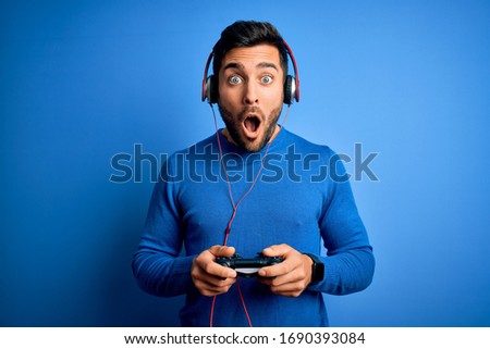 Young handsome gamer man with beard playing video game using joystick and headphones scared in shock with a surprise face, afraid and excited with fear expression