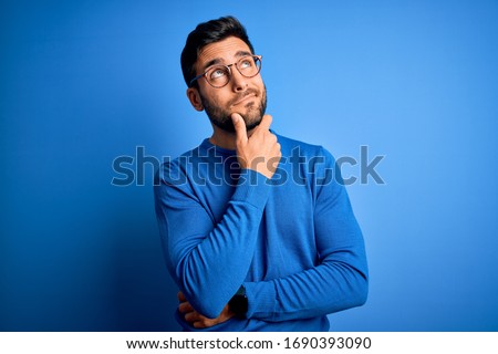 Young handsome man with beard wearing casual sweater and glasses over blue background Thinking worried about a question, concerned and nervous with hand on chin Royalty-Free Stock Photo #1690393090