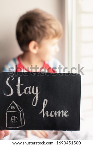 Drawing with chalk on a black blackboard, house with text Stay Home. Child sitting near window on background. Concept of virus protection