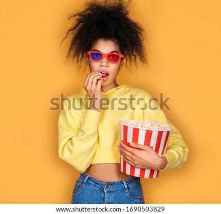 Young girl watching tv with popcorn box. Photo of african american girl on yellow background