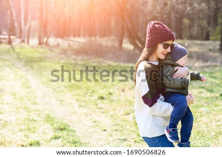 A stylish young mother in a knitted hat and sunglasses carries a baby boy in her arms, they laugh. Family time in the park, in nature, in the forest. Copy paste space for the text on the picture