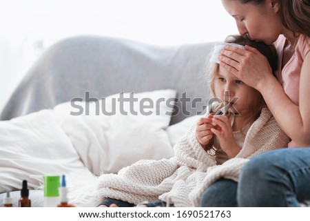Mother checks her daughter's temperature on a thermometer. The little girl is sick and sitting on the sofa, she is ill. A mother cares, hugs and kisses her daughter. There are medicines on the table.