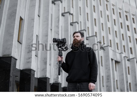 Bearded Professional videographer in black hoodie holding professional camera on 3-axis gimbal stabilizer. Filmmaker making a great video with a professional cinema camera. Cinematographer.
