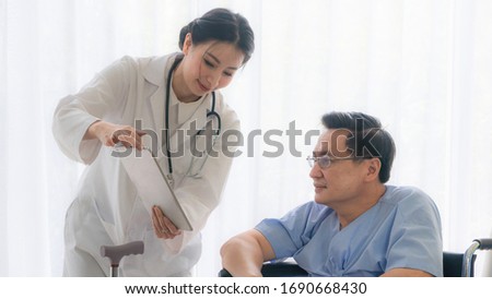 Asian woman doctor gives advice on tablet computer to an elderly old senior patient sit in wheelchair in Interior patient room background white curtain in hospital.