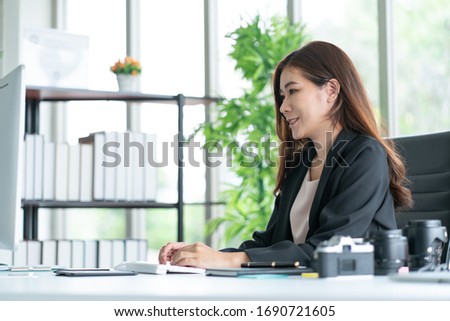 Asian business woman sitting in office looking at computer monitor with smile while camera and lens on her desk. Photography business concept.