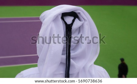 An unidentified Qatari man seen in national costume from behind looking out at a tennis court. It looks as if he is waiting for the sport to resume. In front of him there is a person dressed in black. Royalty-Free Stock Photo #1690738636