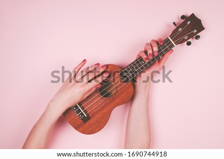 Woman hands with beauty pink manicure holding little wodden ukulele on pink background. Fine female musician picture.