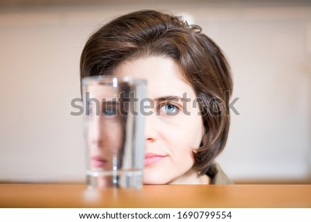 Portrait of beautiful woman. Girl looks in a glass of water on the table. Half-face photo. Creative refraction photography
