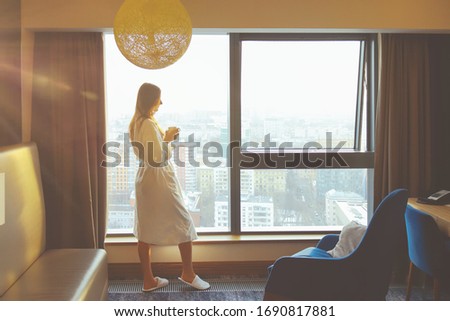 Silhouette of a young beautiful woman with cup of coffee in the white bathrobe on the hotel window background at the yellow sunrise time.