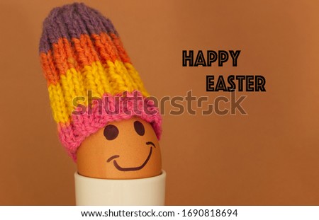 Easter themed photo. Brown egg with a smiling face wearing a bright coloured hat (knitted homemade egg cosy). Happy Easter written on the right hand side and a brown back ground.