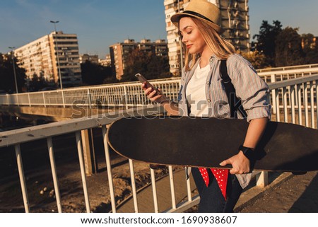 Smiling girl walking in the city carrying long board and phone in her hands.
