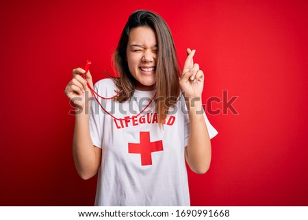Young beautiful brunette lifeguard girl wearing t-shirt with red cross using whistle gesturing finger crossed smiling with hope and eyes closed. Luck and superstitious concept.