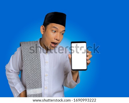 Photo image portrait religious asian muslim man in traditional dress isolated over blue background. Showing a mobile phone