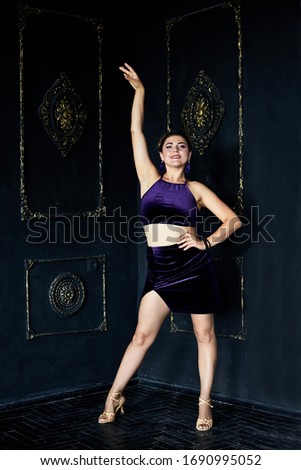 Young flexible brunette woman, wearing dark violet top and skirt, stretching, doing dance posing, in dark loft living room by black wall. Salsa ballet dancer female photo shoot in dark dance studio