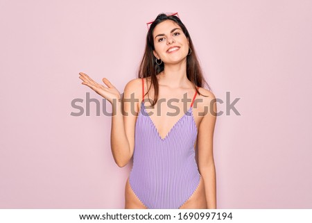 Young beautiful fashion girl wearing swimwear swimsuit and sunglasses over pink background smiling cheerful presenting and pointing with palm of hand looking at the camera.