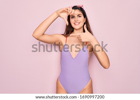 Young beautiful fashion girl wearing swimwear swimsuit and sunglasses over pink background smiling making frame with hands and fingers with happy face. Creativity and photography concept.