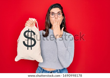 Young beautiful girl holding money bag with dollar symbol for business wealth over red background cover mouth with hand shocked with shame for mistake, expression of fear, scared in silence, secret Royalty-Free Stock Photo #1690998178