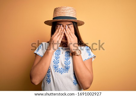 Young beautiful asian girl wearing casual t-shirt and hat standing over yellow background rubbing eyes for fatigue and headache, sleepy and tired expression. Vision problem