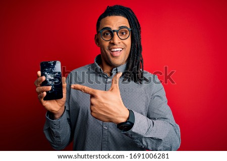 Young african american afro man holding broken smartphone showing craked screen very happy pointing with hand and finger